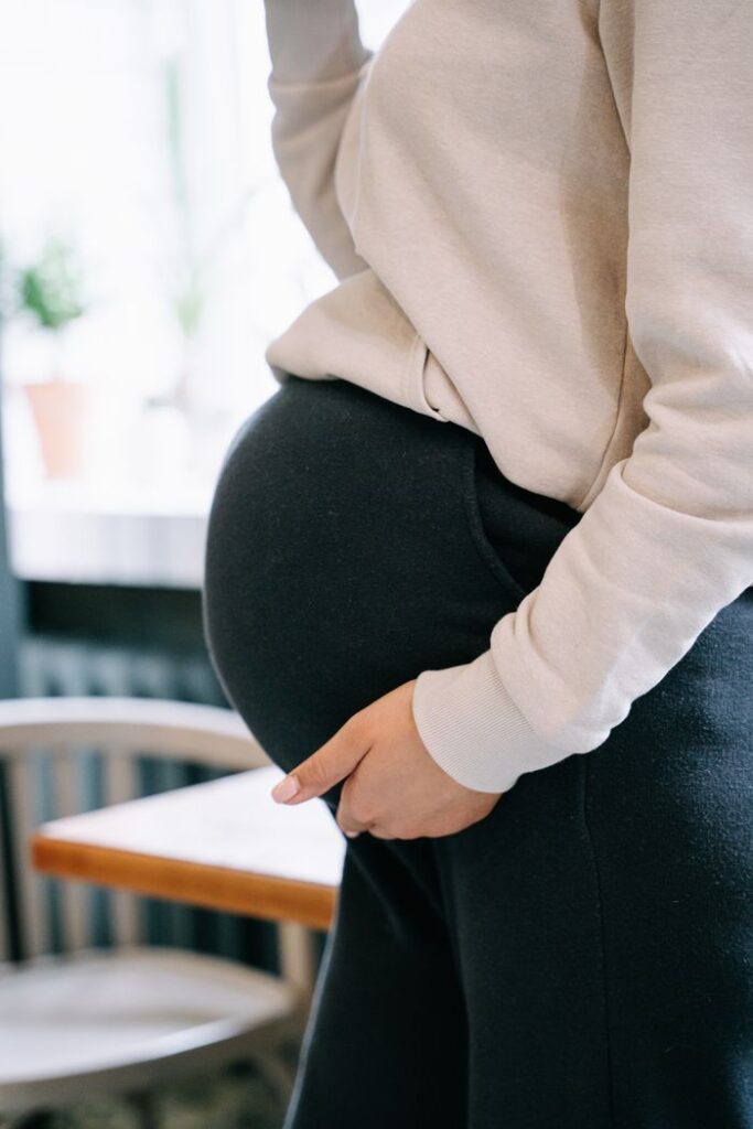 Side profile of a pregnant woman gently holding her belly wearing a white sweater and black pants.